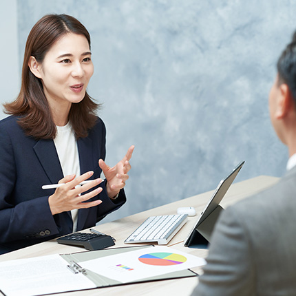 会話をしているビジネスパーソンの女性と男性、デスクに並んでいる資料とパソコンが写っているシーン