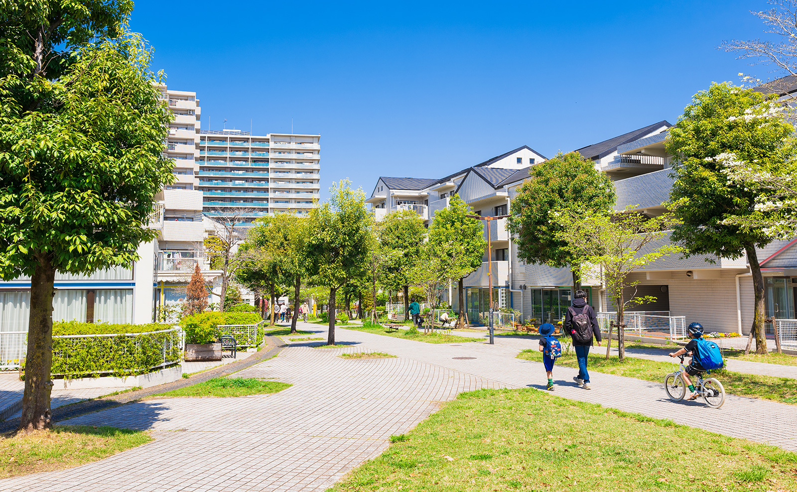 広がる緑地に囲まれたマンションと遊歩道。家族が散歩しているシーン、晴れ渡る青空が広がる都市開発エリアのイメージ